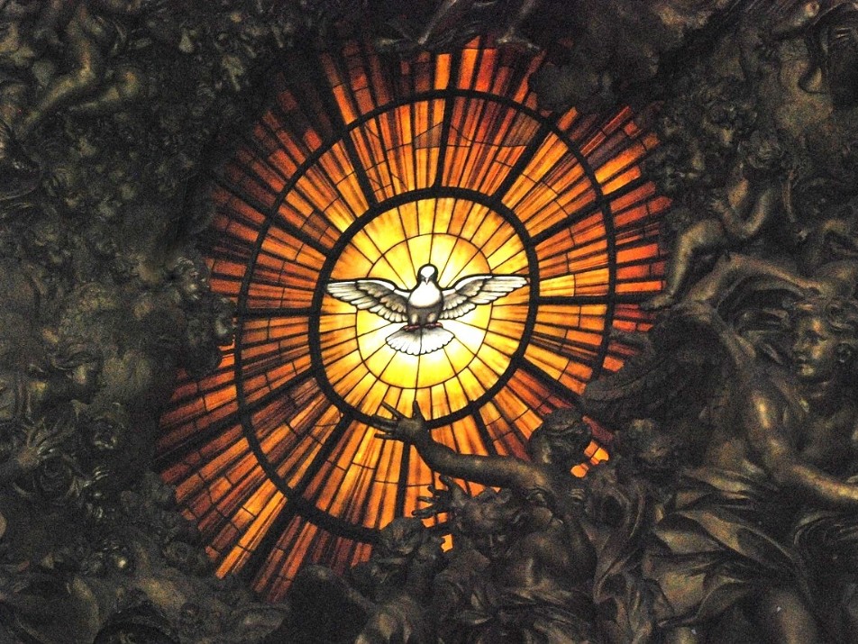 Das Bild zeigt ein rundes  Glasfenster im Petersdom. In der Mitte ist eine weiße Taube, die nach allen Seiten hin ausstrahlt. Zum Rand hin gehen die Farben von Weiß und Gelb in Orange über und werden dunkler. Das Fenster ist umrahmt von vielen Gestalten, die im Dunklen sind. Die Taube - Symbol für Gottes Geist des Friedens - strahlt nach allen Seiten aus, bis in die Dunkelheit der Welt.